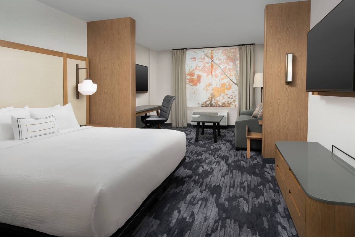 A hotel room with a large bed, a desk, a chair, a TV, and a window with a view of trees.