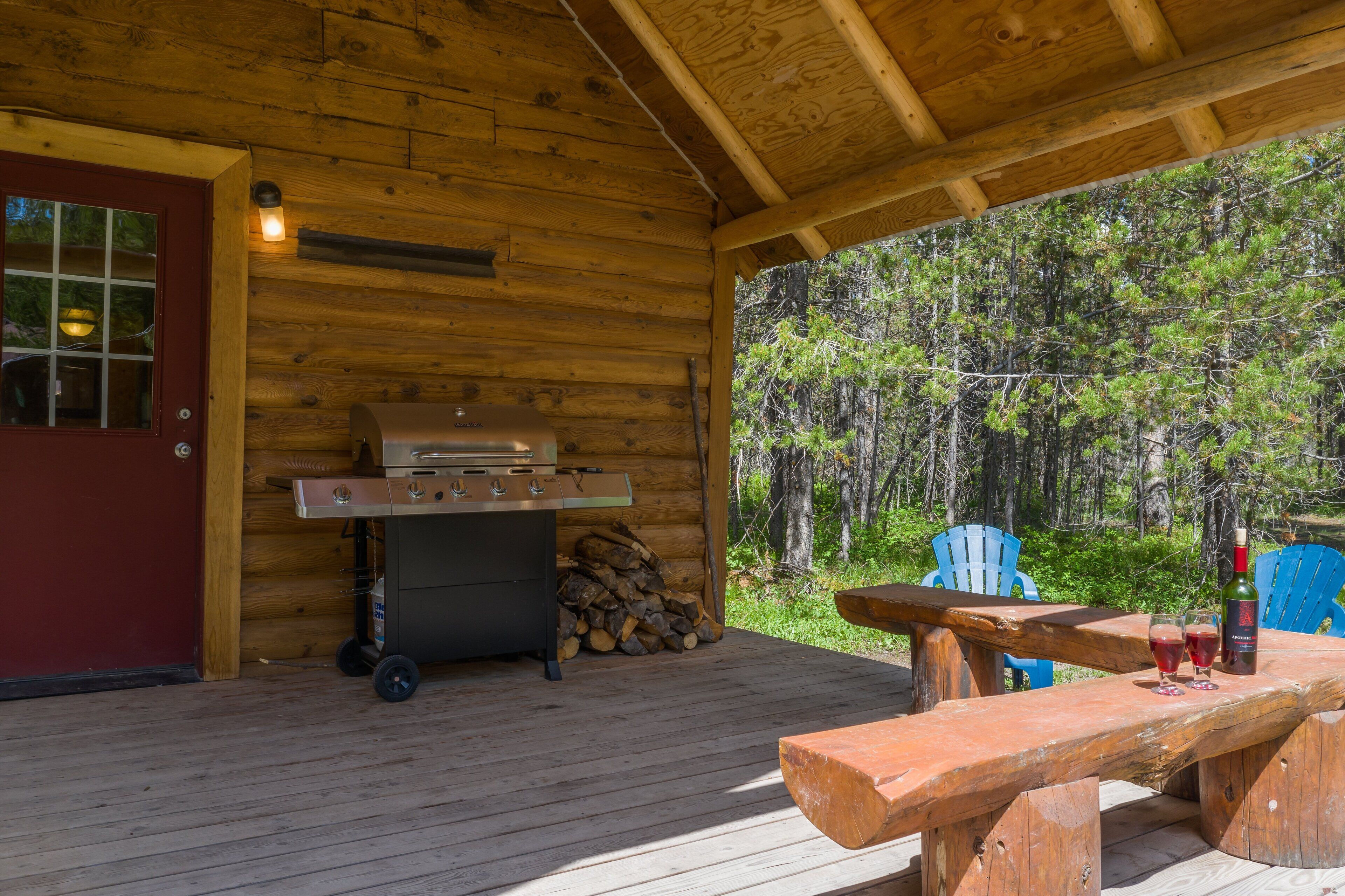 A wooden cabin with a barbecue grill, a red door, and a picnic table with glasses and a bottle.