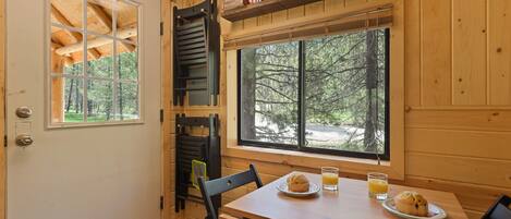 Interior de una cabaña de madera con una mesa de comedor, sillas, una ventana que ofrece vistas a árboles y una estantería con cajas.