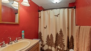 A bathroom with a red wall, a beige sink, a mirror, and a shower curtain with a forest design. - Little Fox (Island Park)