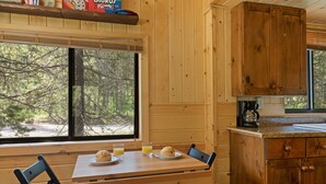 A wooden cabin interior with a dining table set for breakfast, a coffee maker, and a window overlooking a forest. - Little Fox (Island Park)