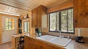 Une cuisine rustique, située dans une cabane en bois, avec un évier à deux bacs, des placards en bois et une fenêtre donnant sur une forêt.