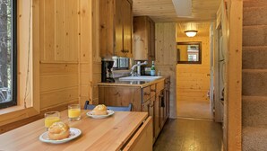 L’intérieur d’un chalet en bois comprend un coin cuisine, une table à manger avec des muffins et du jus, et un escalier menant à une autre pièce.