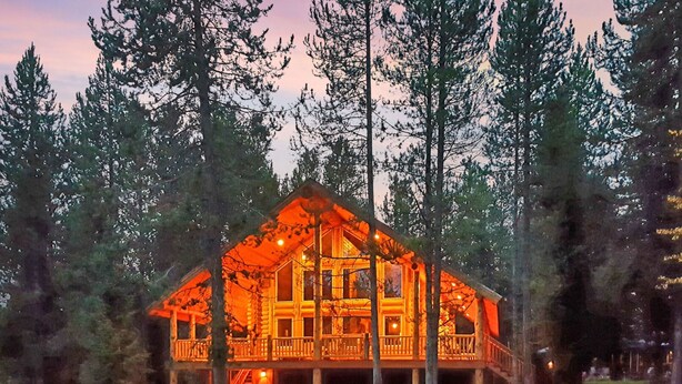 A log cabin with a porch and a staircase leading to the entrance, surrounded by tall trees. - Lumberjack Lodge (Island Park)