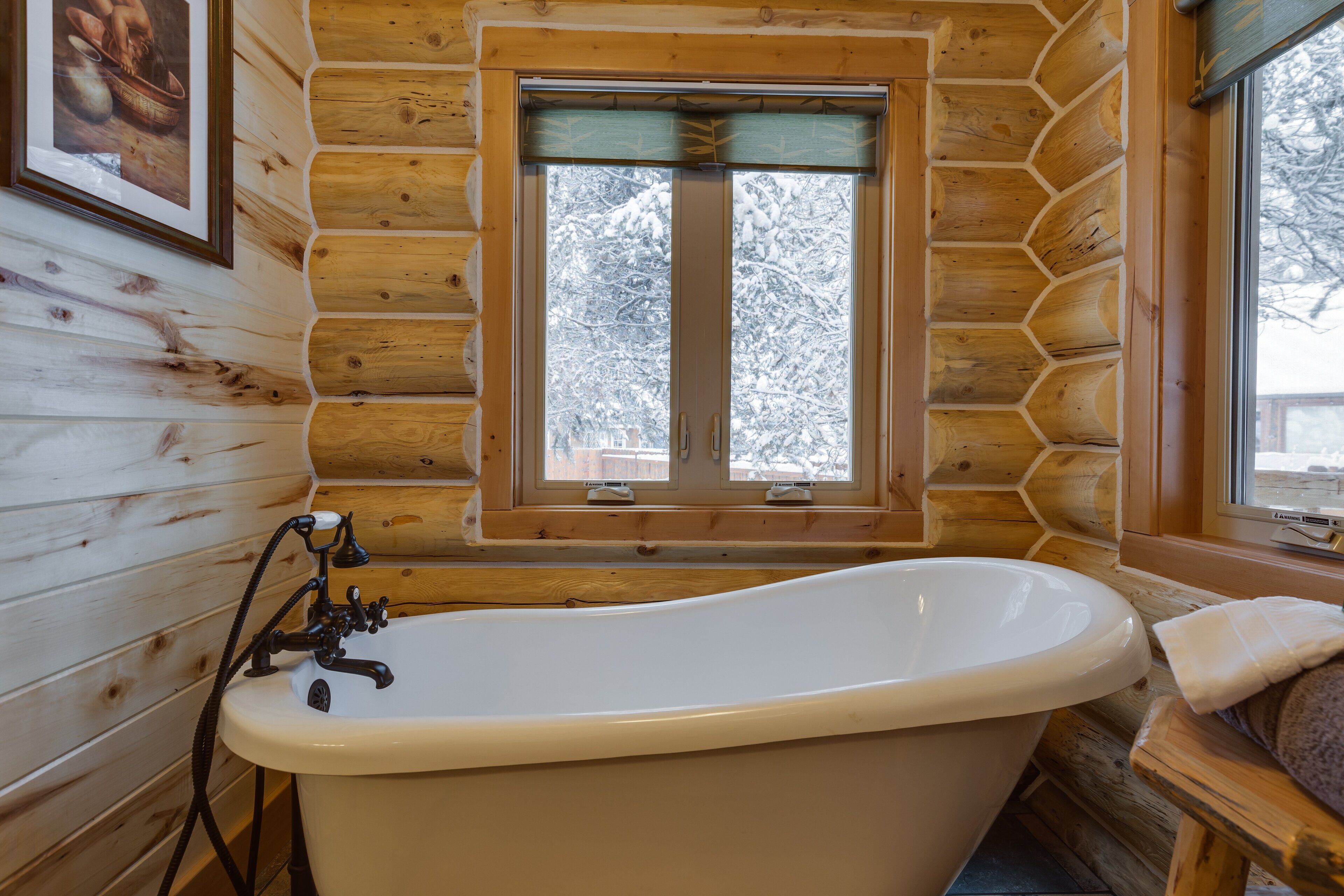 Ein Badezimmer in einer Blockhütte mit freistehender Badewanne, Holzwänden und einem Fenster mit Blick auf schneebedeckte Bäume.
