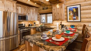 A modern kitchen with granite countertops, stainless steel appliances, wooden cabinetry, and a dining area with a long table set for four. - Lumberjack Lodge (Island Park)