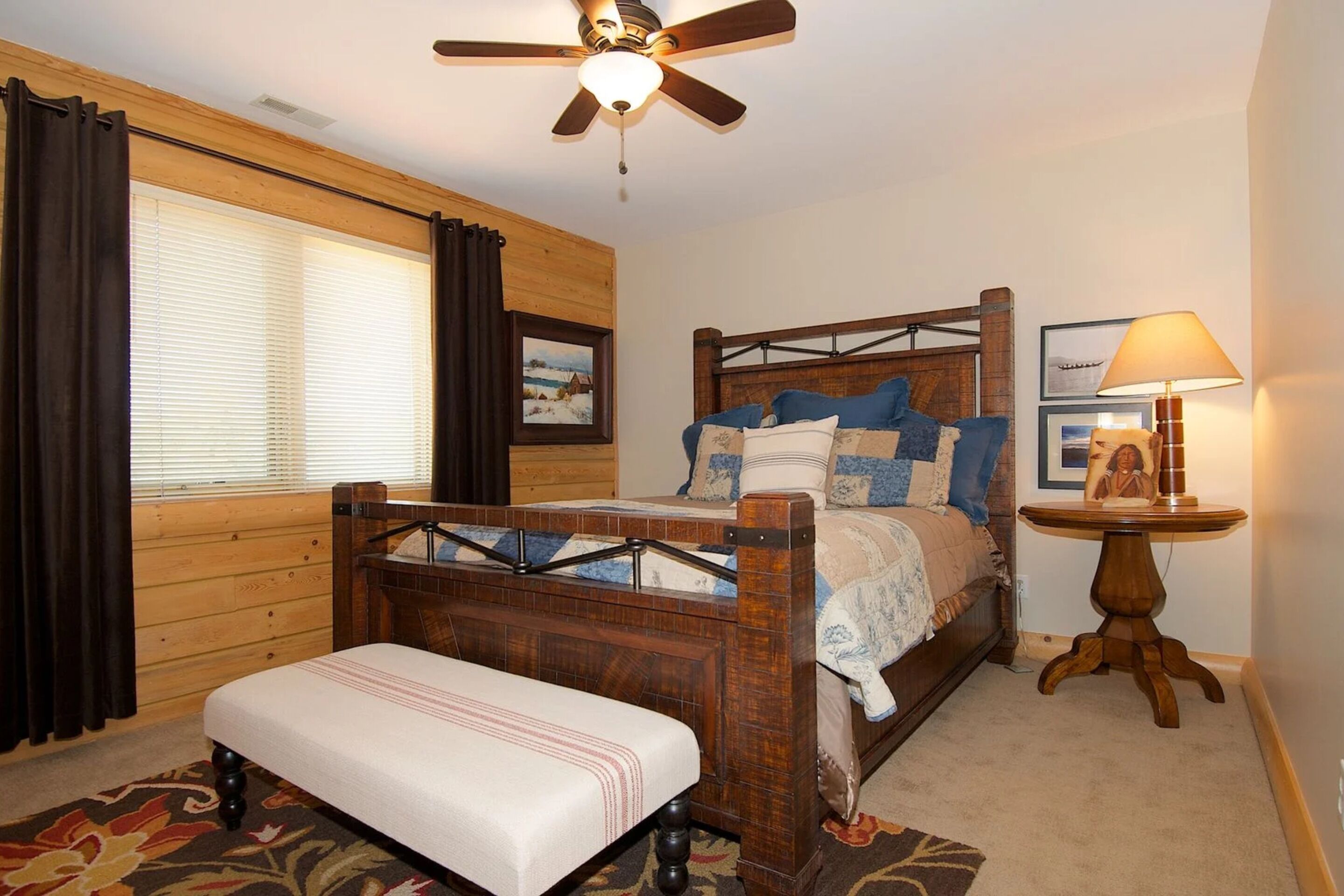 A bedroom with a wooden bed, a bench, a ceiling fan, a window with curtains, and a small table with a lamp.