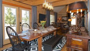 A wooden dining room with a long table set for four, surrounded by chairs. There is a bench at the end of the table, and a chandelier hangs above. - Eagles Perch (Island Park)