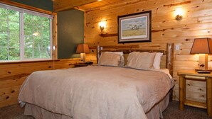 A wooden cabin bedroom with a large bed, two bedside tables with lamps, a framed picture on the wall, and a window with a view of trees.