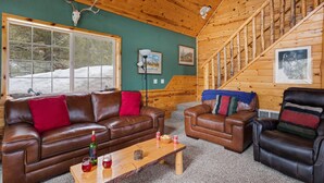A cozy cabin interior with a wooden staircase, leather sofas, and a large window overlooking a snowy landscape.