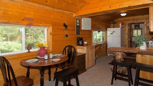 A wooden cabin interior with a dining area, kitchen, and a large window overlooking greenery. - Remington Retreat (Island Park)