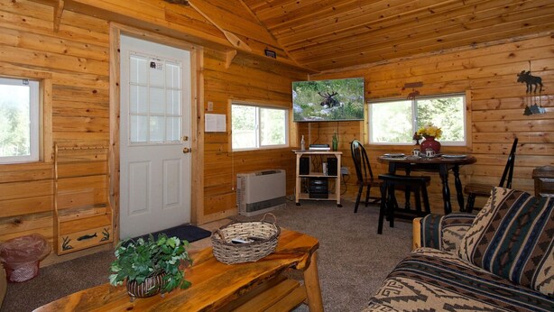 A log cabin interior with a wooden door, a television mounted on the wall, a dining table with chairs, and a sofa. - Remington Retreat (Island Park)