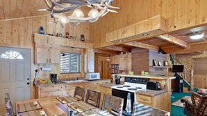 A wooden cabin interior with a dining area, kitchen, and a staircase leading to a loft. - Restoration Pines (Island Park)