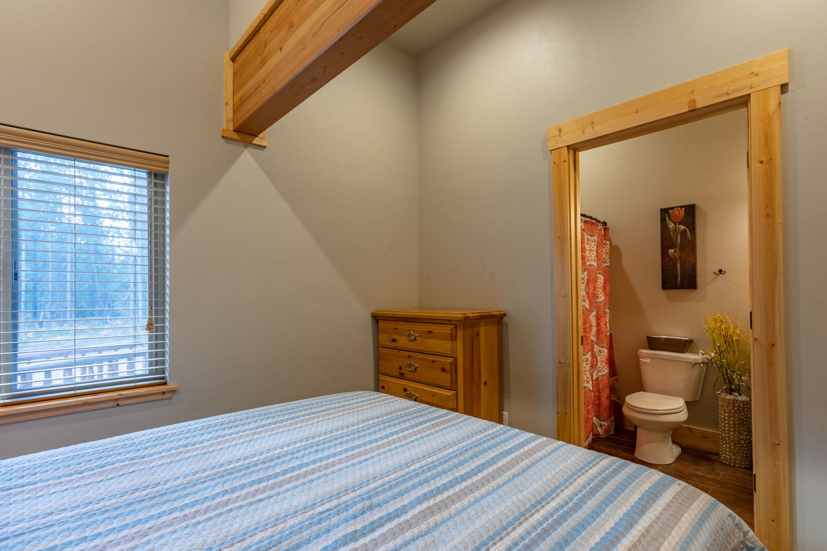 Une chambre à coucher avec un lit, une commode en bois, une fenêtre avec des stores et une salle de bain visible à travers une porte ouverte.