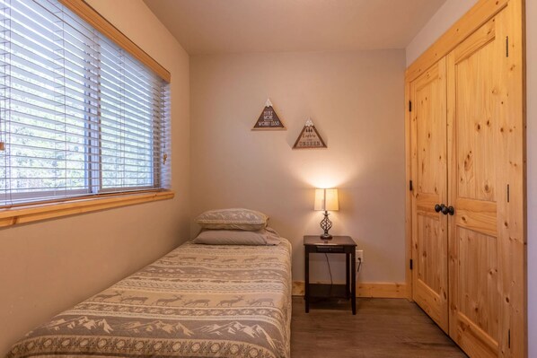 A bedroom with a bed, a bedside table with a lamp, a window with blinds, and a wooden wardrobe. - Buck Buck Moose (Island Park)