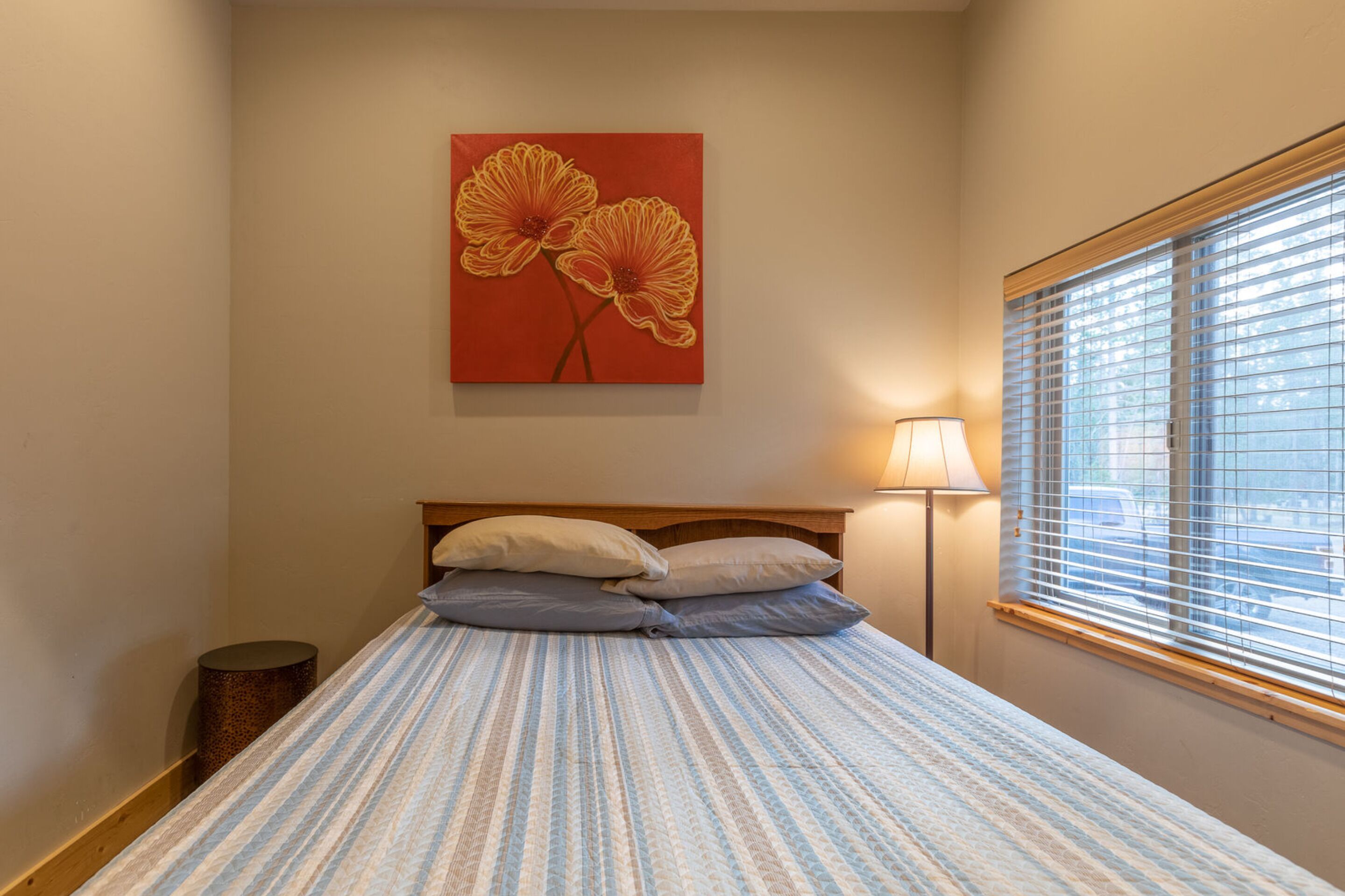 A bedroom with a bed, pillows, a lamp, a window with blinds, and a painting on the wall.