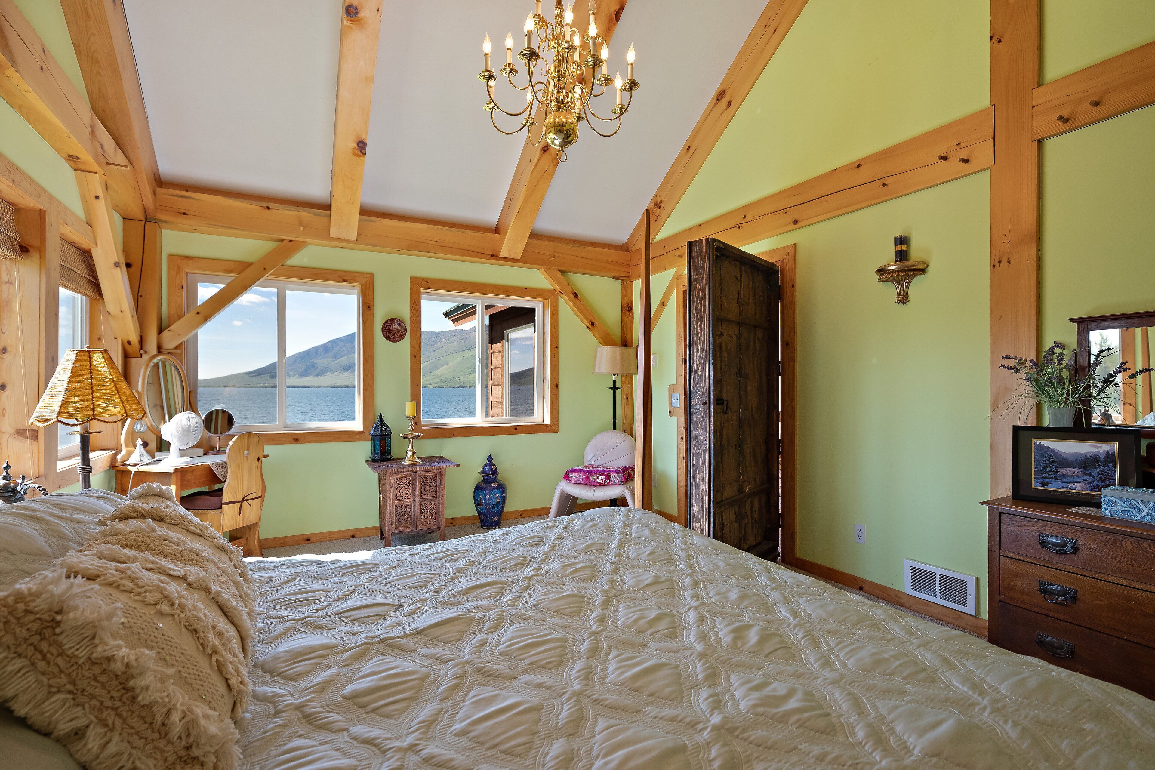 A bedroom with a large bed, wooden beams, a chandelier, a dresser, and a window with a view of the outdoors.