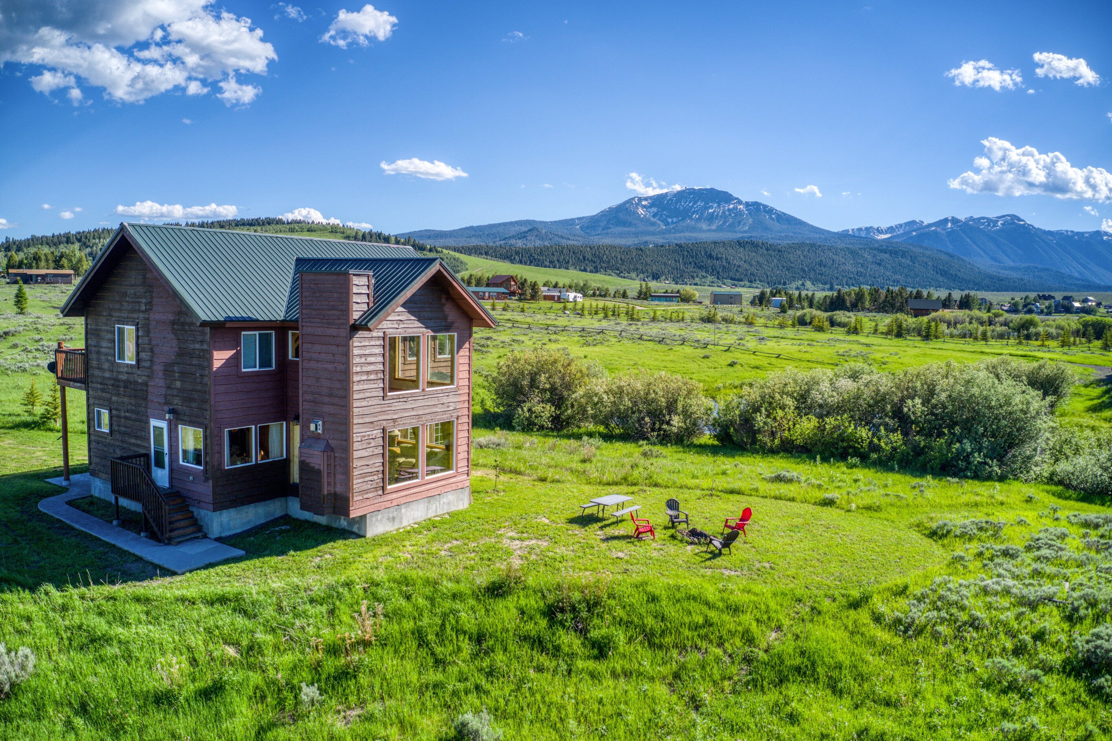 Une cabane en rondins à deux étages, surmontée d’un toit en métal vert, située dans un champ verdoyant, avec des montagnes en toile de fond.