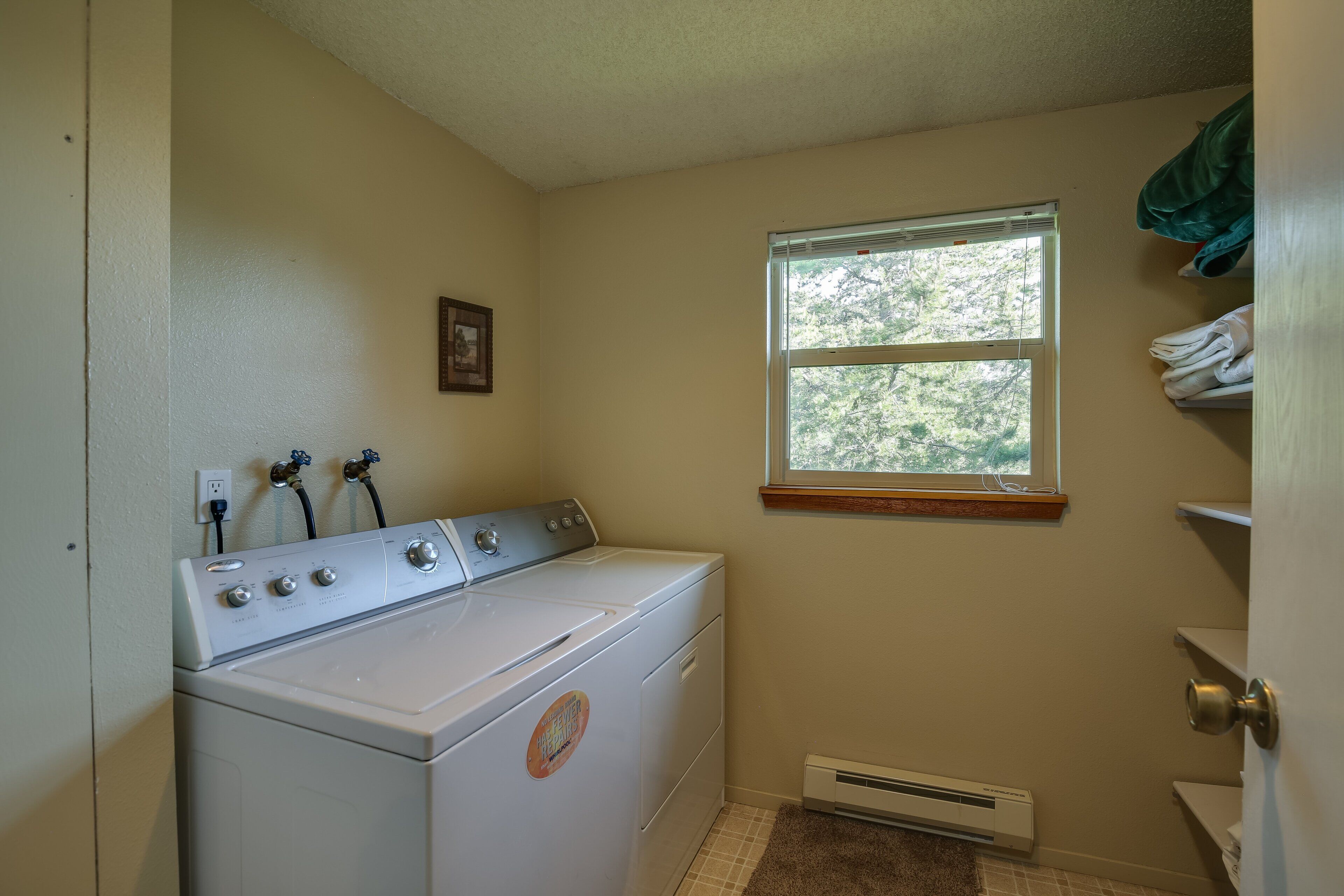 A laundry room with a front-loading washer and dryer, a window with a view of trees, and a wall-mounted heater.