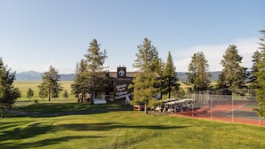 En klubblokal med en skylt som lyder ”Island Park Village Clubhouse”, omgiven av en tennisbana med staket och en gräsyta med träd.