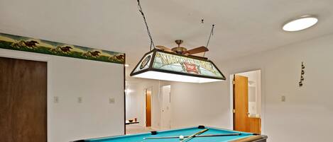 A pool table with a wooden frame and a green felt surface, a ceiling fan, and a wall-mounted light fixture.