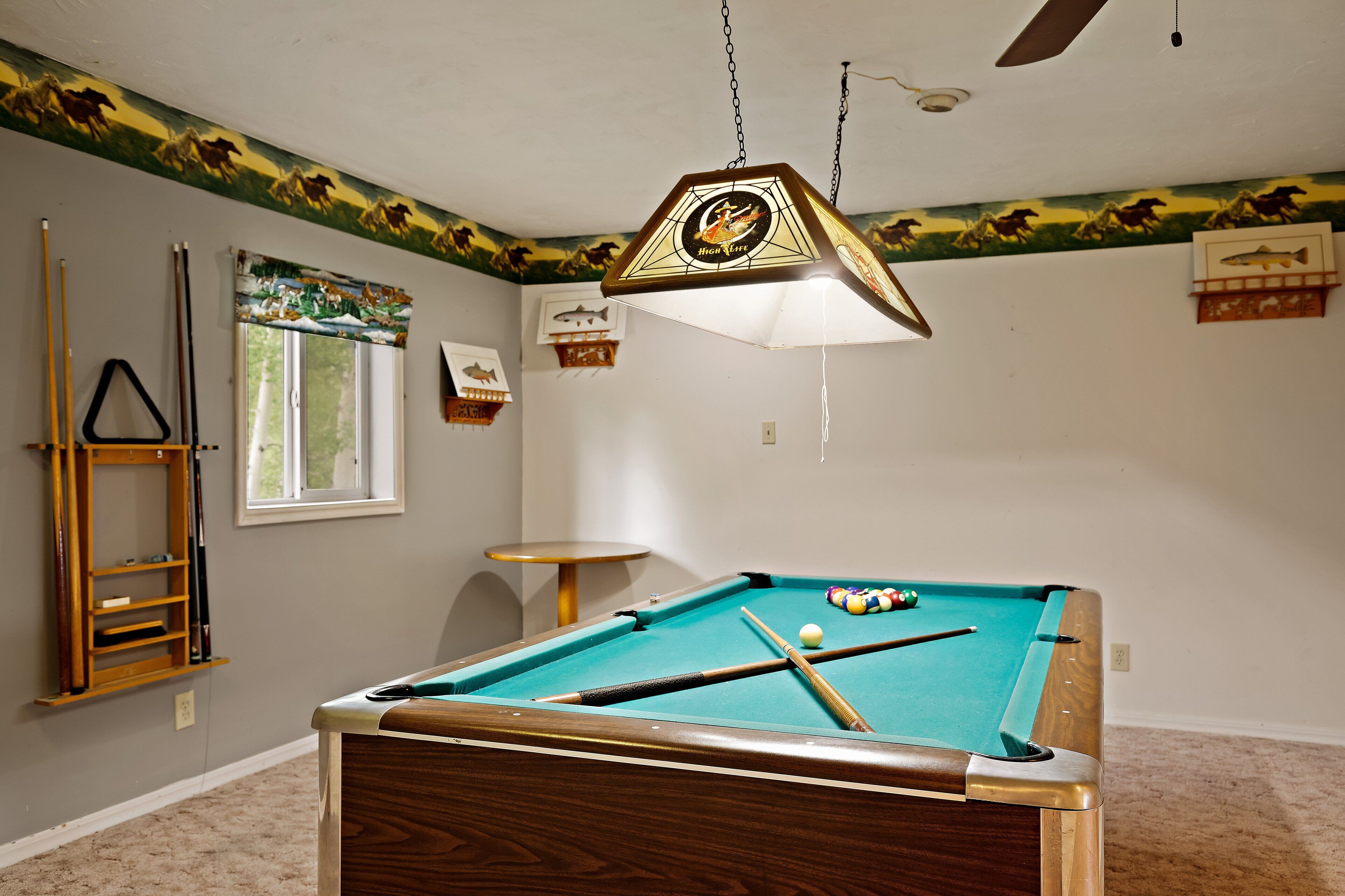 A pool table with a wooden frame, a hanging light fixture, and a window with a view of the outdoors.