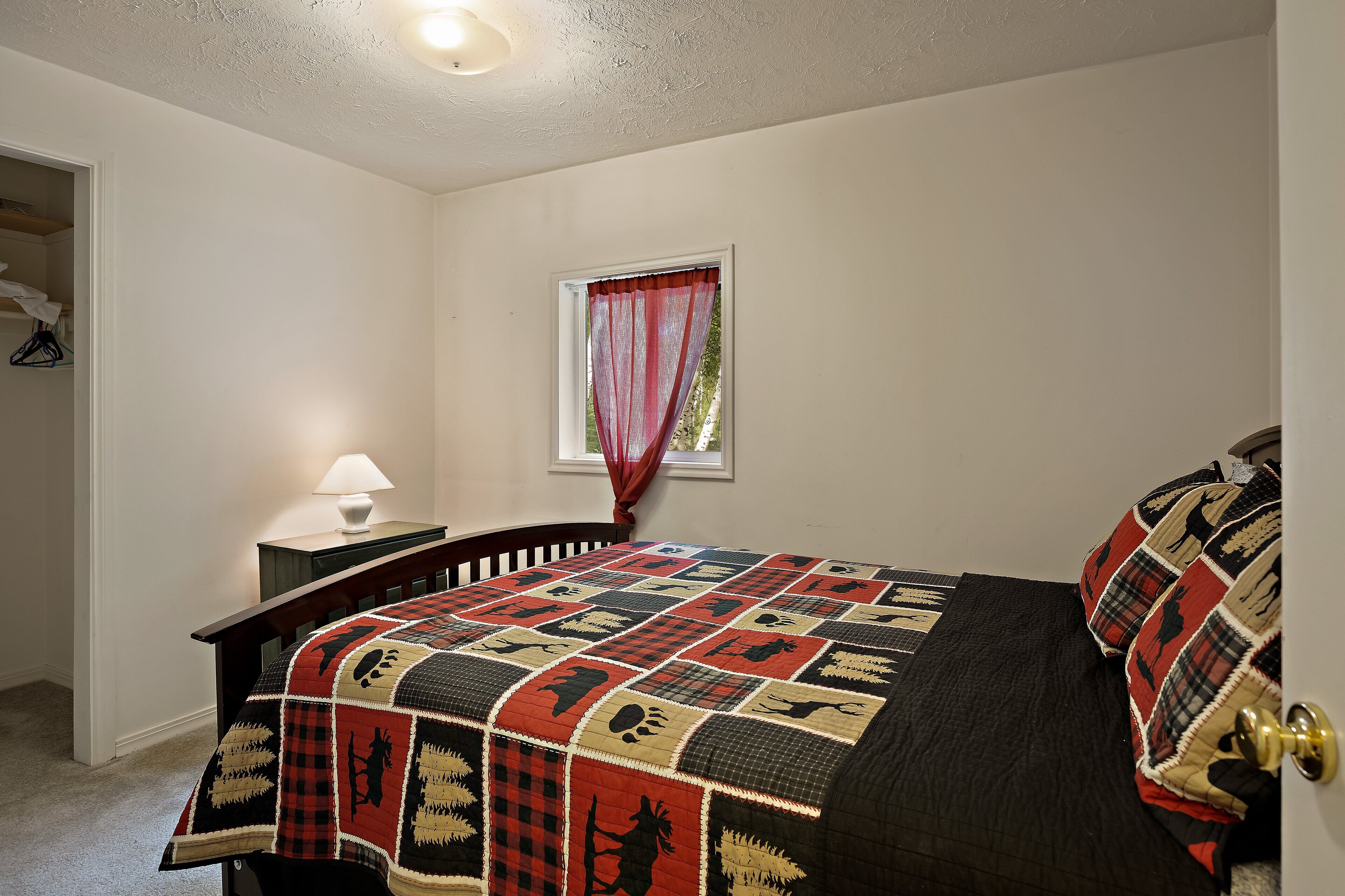 A bedroom with a large bed, a nightstand with a lamp, a window with a red curtain, and a door with a brass handle.