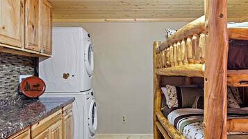Habitación compacta con lavadora, cama con cabecera de madera y zona de cocina con armarios de madera y encimera de mármol.