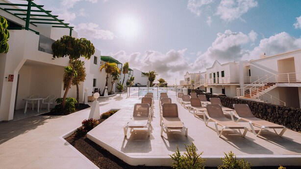 A sunlit outdoor lounge area with rows of sun loungers and a pool in the background.