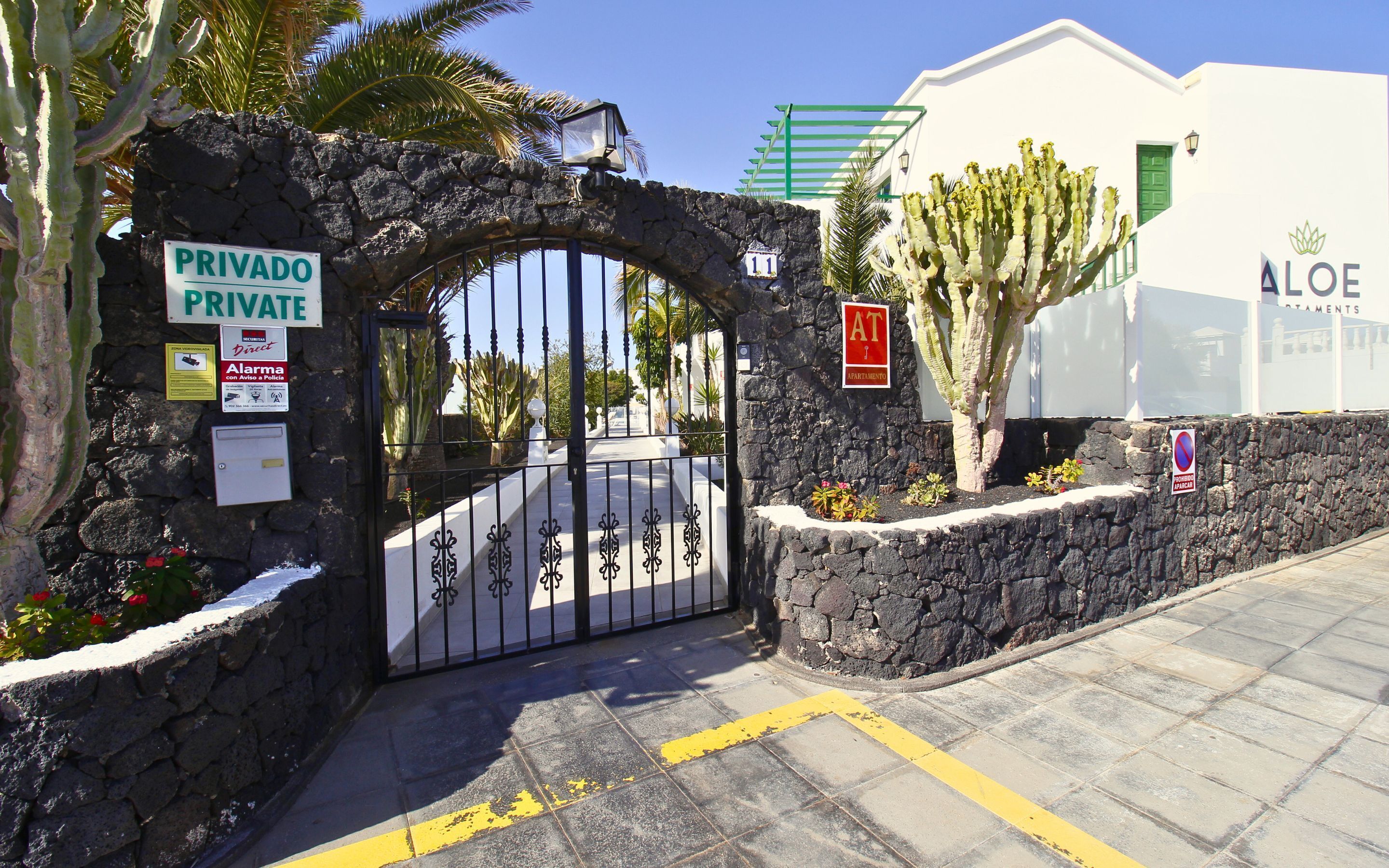 Une porte d’entrée privée avec un mur en pierre, une pancarte indiquant « Privado » et « Alarme avec notification à la police », et un bâtiment portant l’inscription « Aloe ».