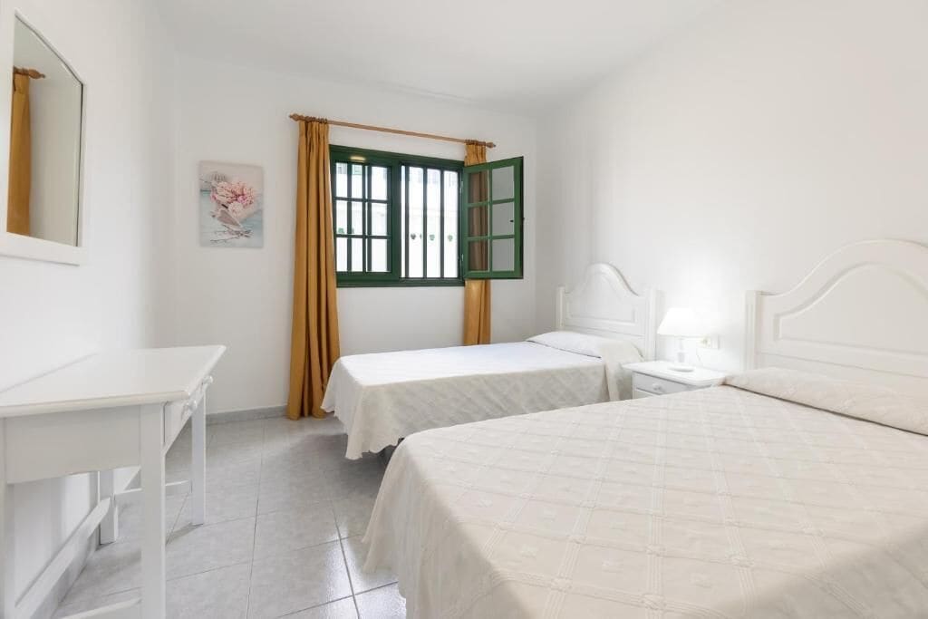 A room with two single beds, a white desk, a chair, a nightstand with a lamp, and a window with green shutters.