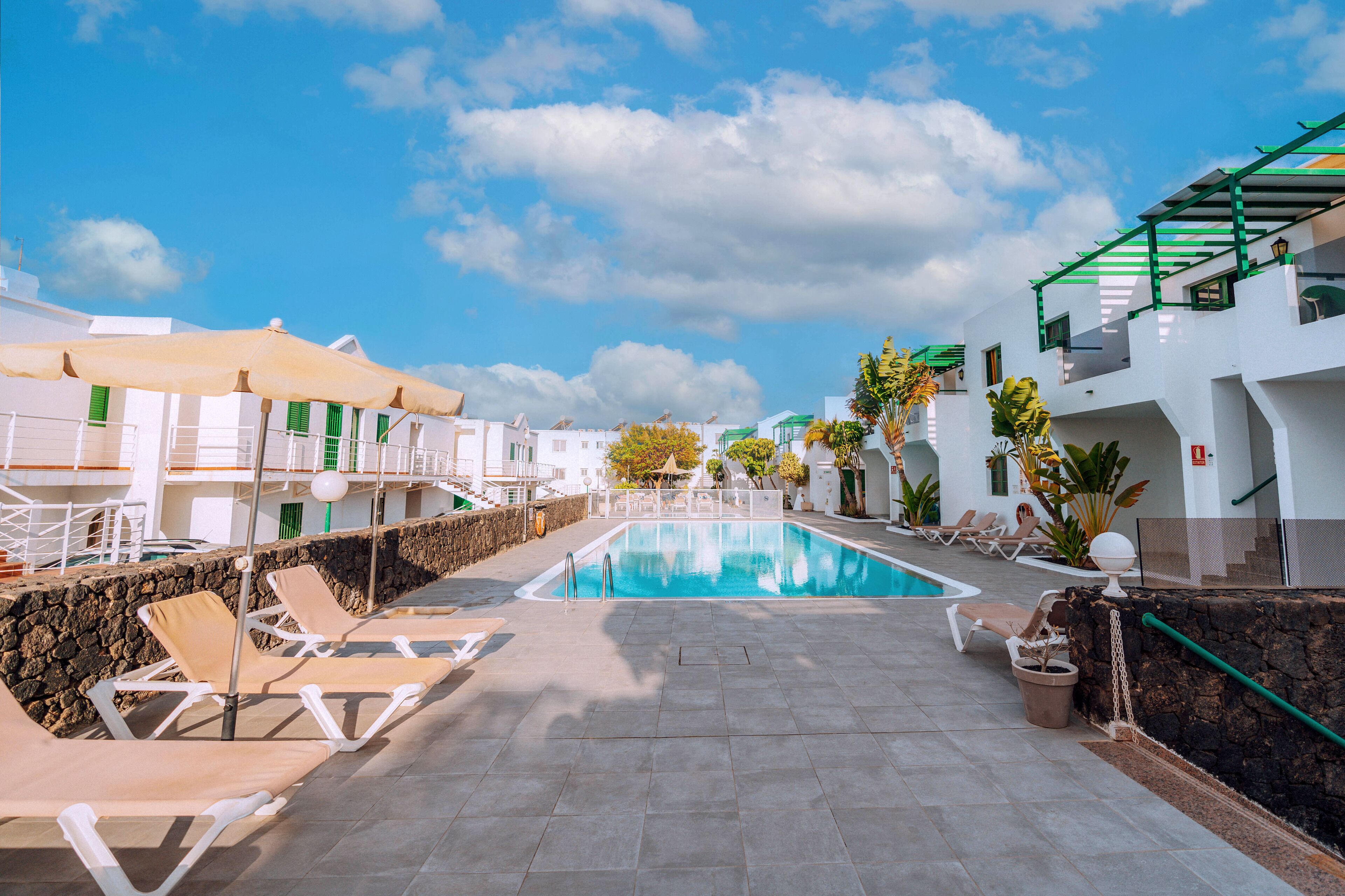Un área de piscina con sillones de descanso, una piscina azul cristalina y edificios blancos con contraventanas verdes.