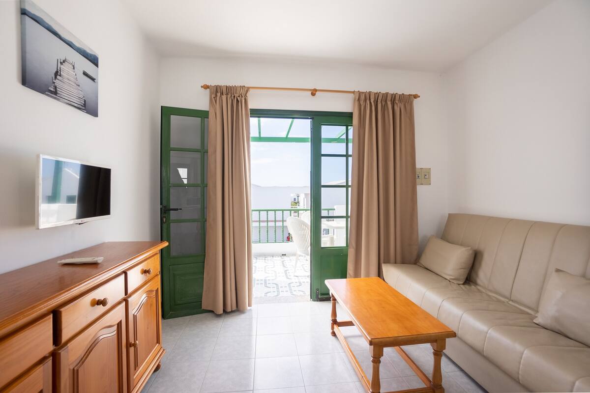 Un salon avec un canapé, une table en bois, un téléviseur posé sur un meuble en bois et une porte verte donnant sur un balcon.