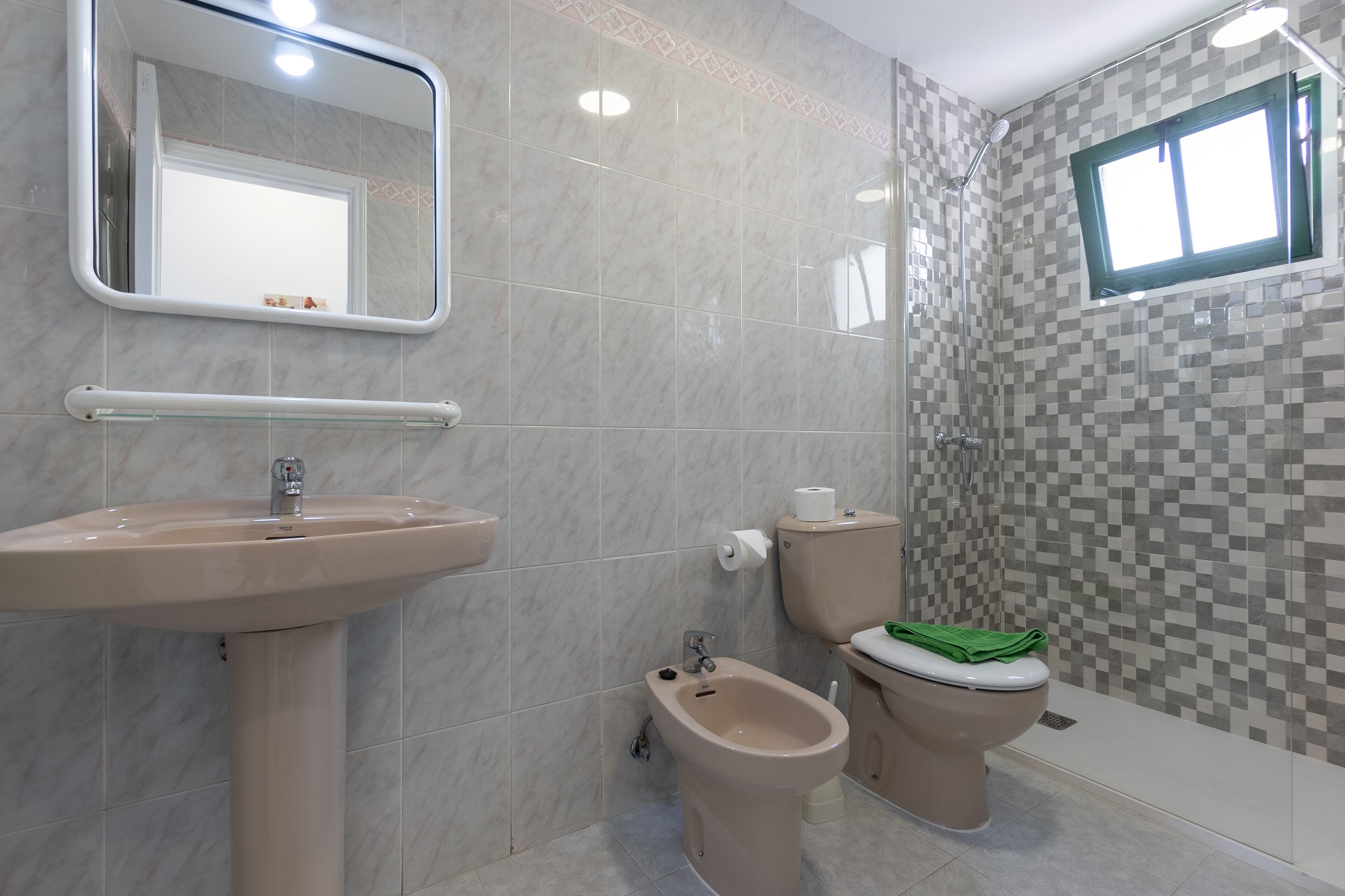 Un baño con lavamanos pedestal, un bidé, inodoro con una toalla verde, espejo y ducha con pared de azulejos.