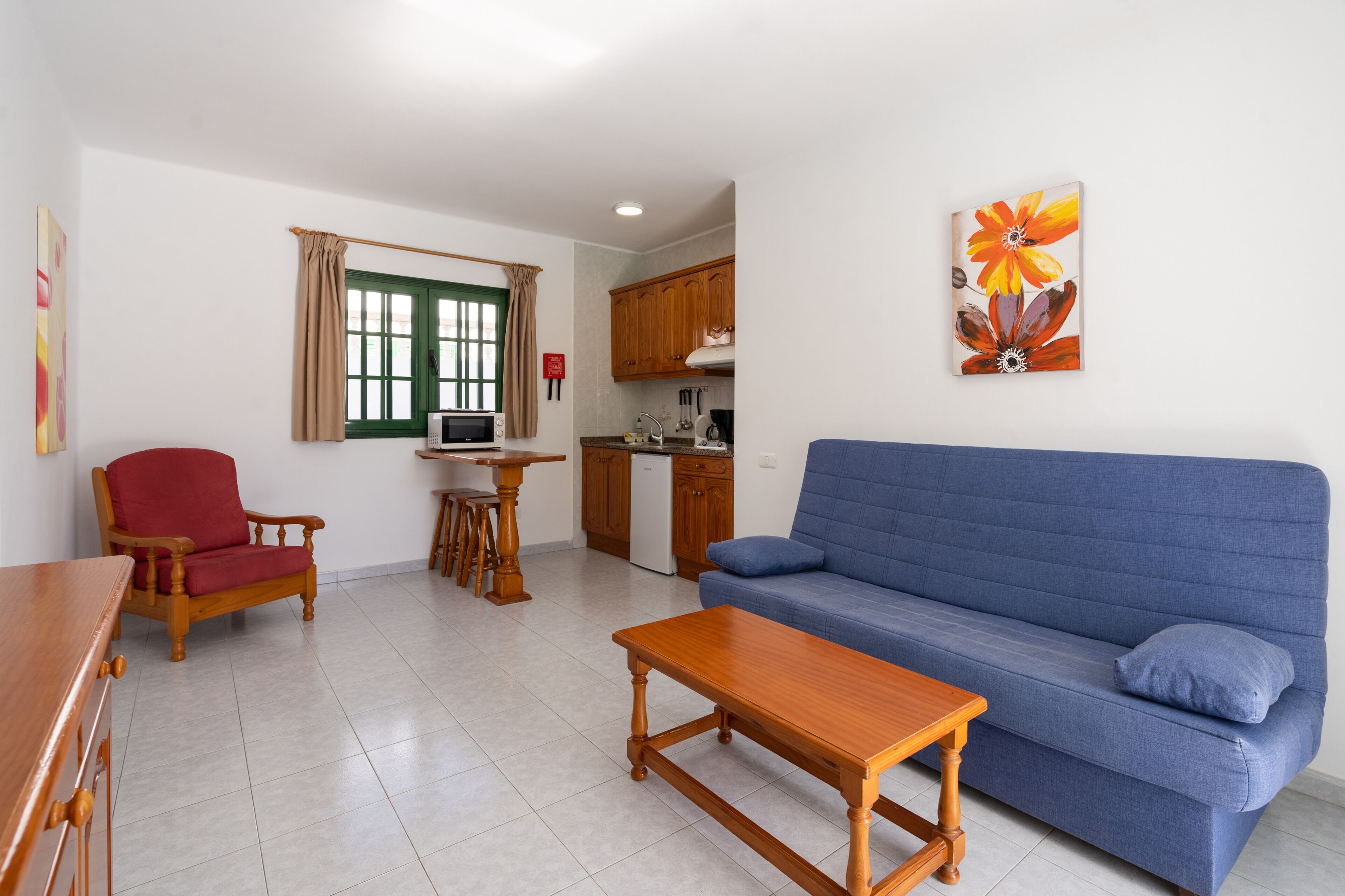 A living room with a blue sofa, wooden coffee table, and a red chair. There is a kitchen area with wooden cabinets and a microwave. A painting of flowers hangs on the wall.