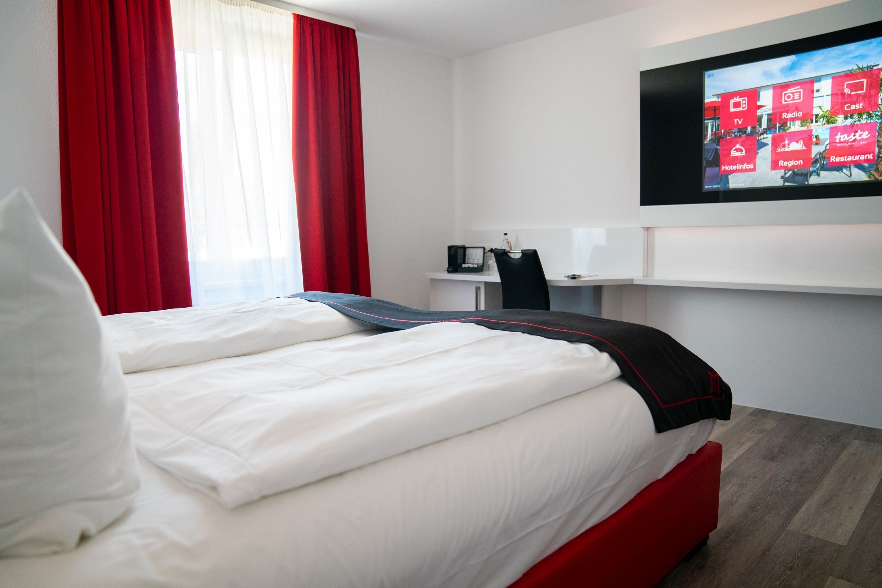 A hotel room with a bed, a desk, a TV, and red curtains.