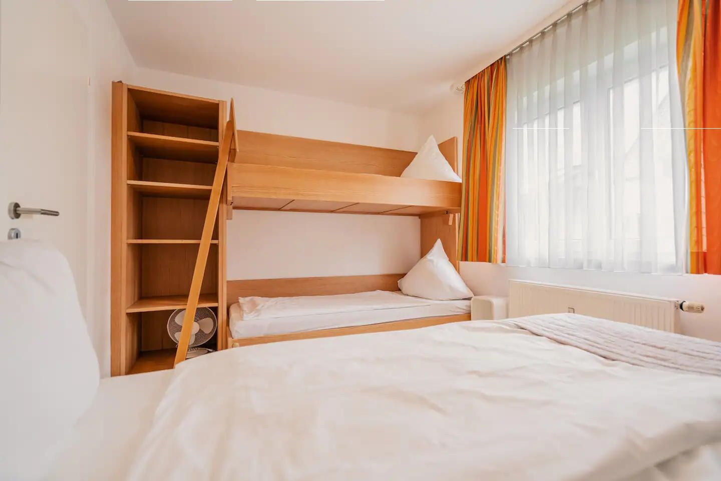 A bunk bed room with a wooden headboard, a small bedside table, and a window with sheer curtains.