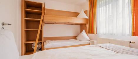 A bunk bed room with a wooden headboard, a small bedside table, and a window with sheer curtains.