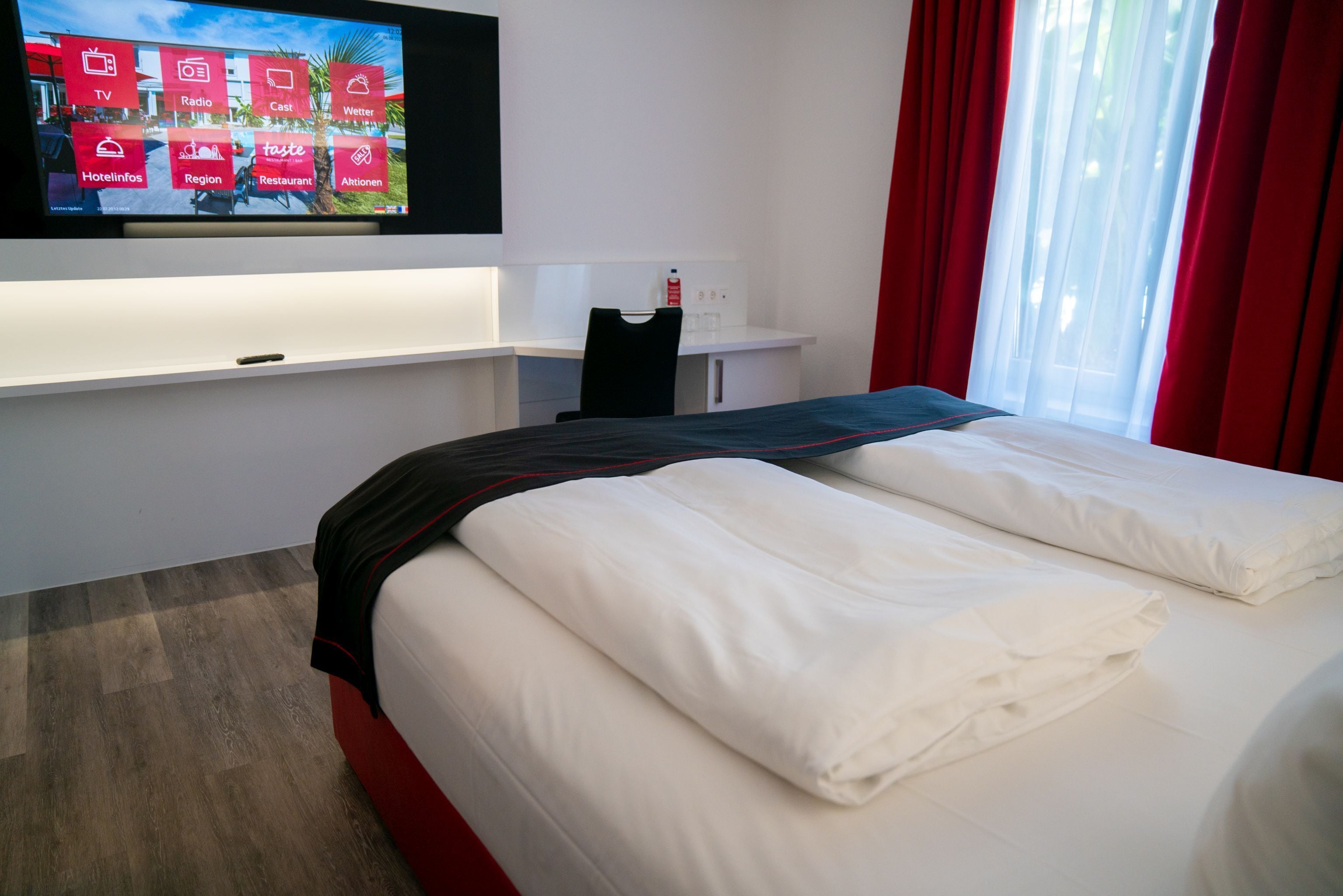 A hotel room with a bed, a TV mounted on the wall displaying hotel amenities, a desk with a chair, and a window with red curtains.