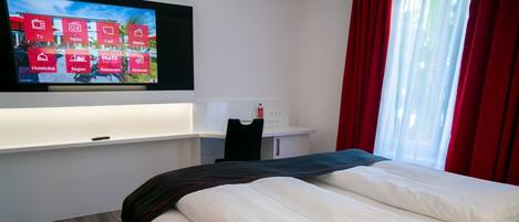 A hotel room with a bed, a TV mounted on the wall displaying hotel amenities, a desk with a chair, and a window with red curtains.
