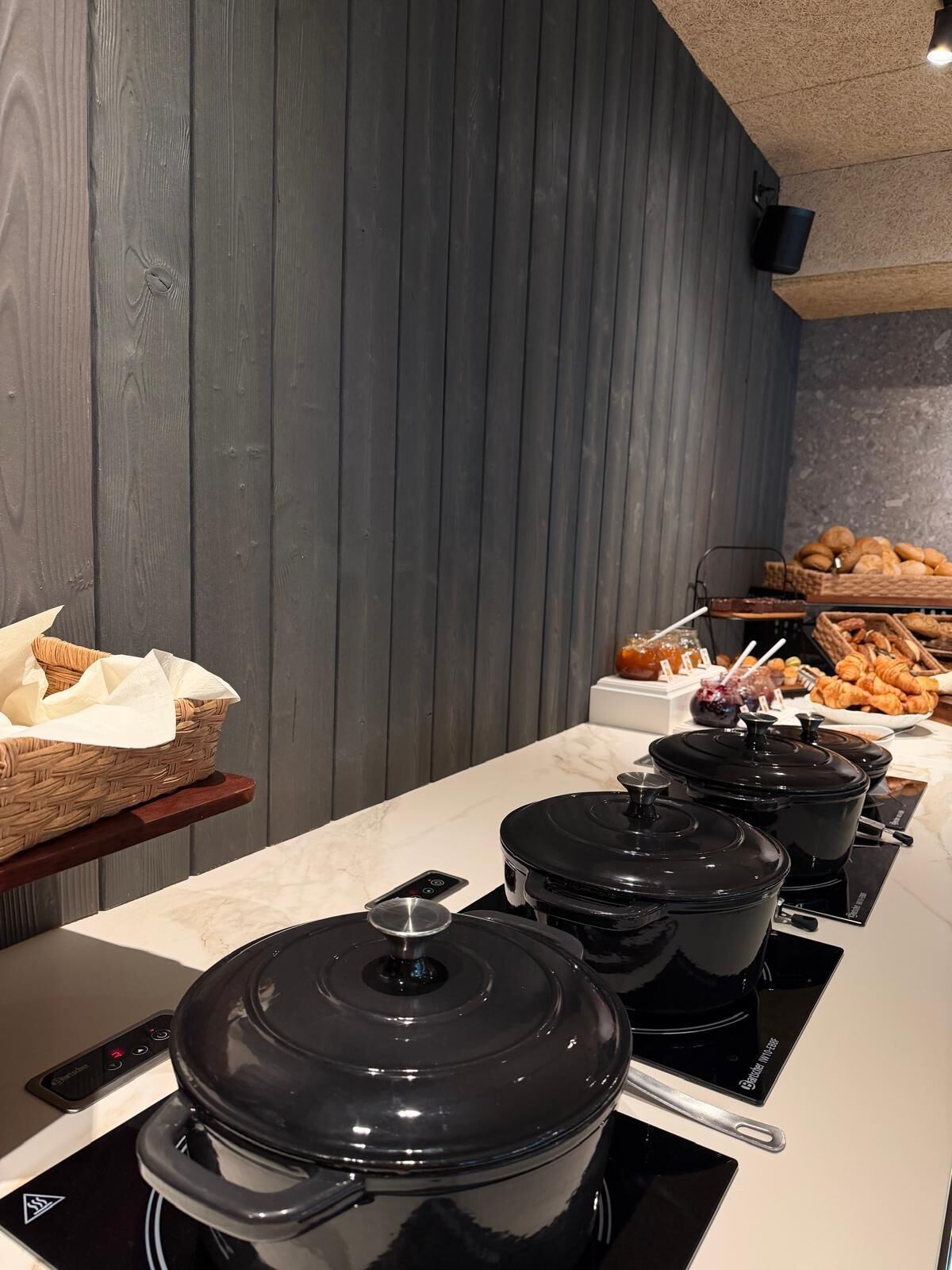 A buffet setup with multiple black pots on induction cooktops, baskets of bread, and a variety of pastries.