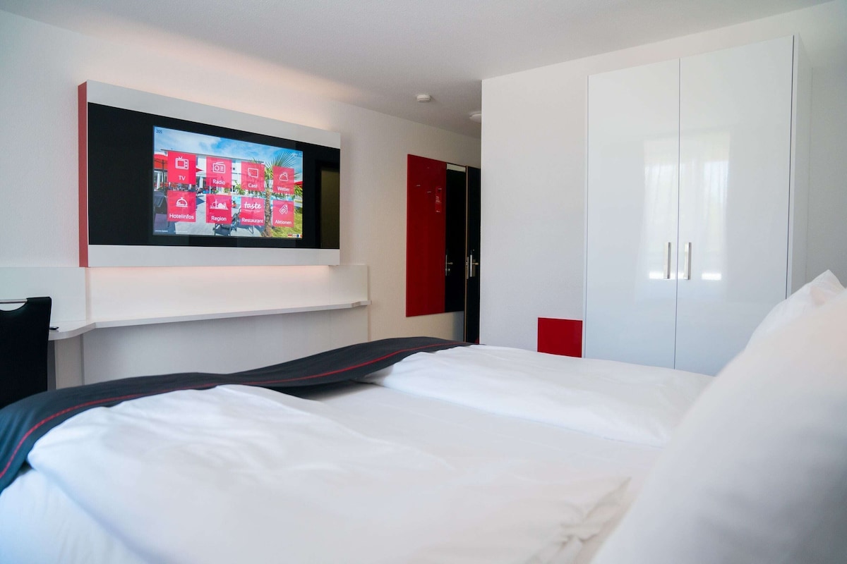 Hotellihuone, jossa on sänky, seinälle asennettu televisio, vaatekaappi ja ovi.