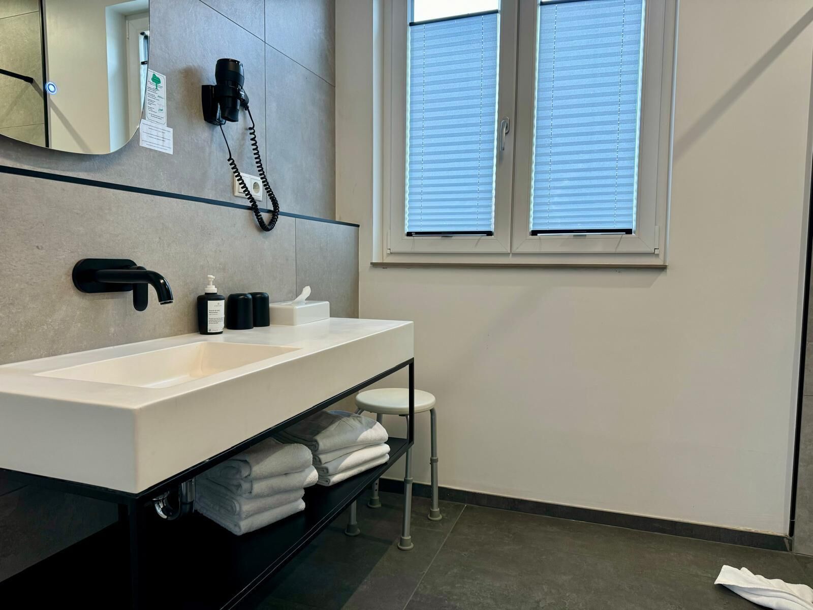 Un baño moderno con un lavabo blanco, grifo negro y un espejo encima. Se ve un perchero con toallas dobladas, un taburete blanco pequeño y una ventana con persianas.
