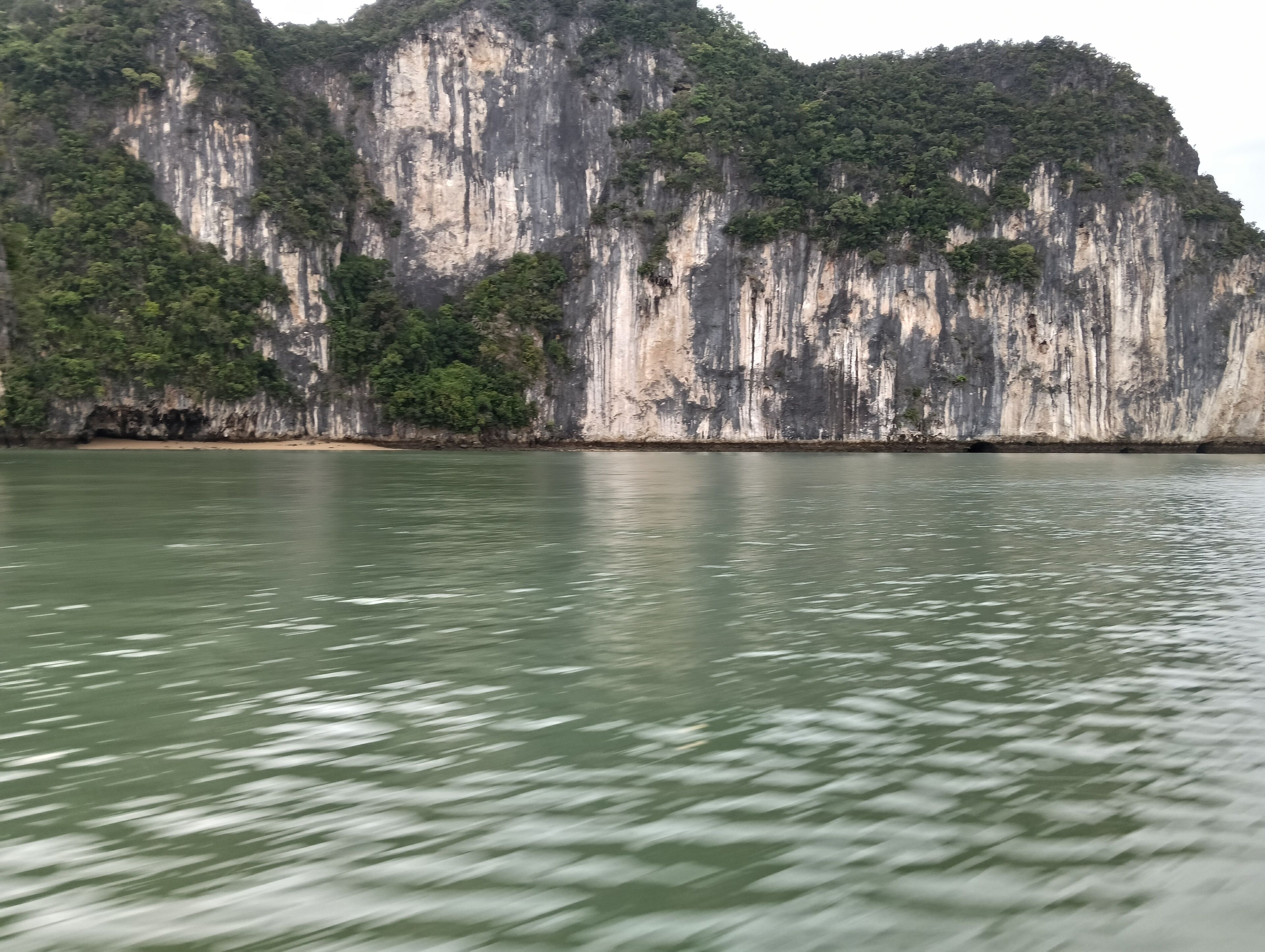 Uma grande formação rochosa natural coberta por vegetação densa, com um corpo d'água calmo ao primeiro plano e uma praia de areia na base.