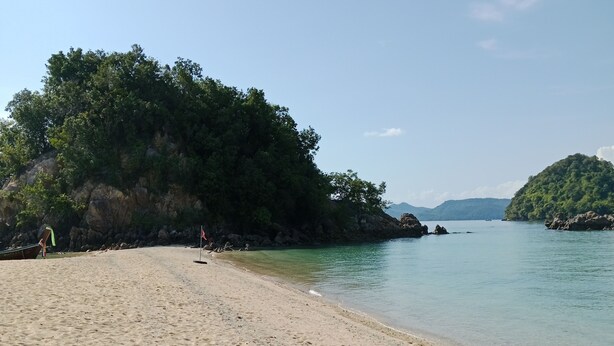 Ein sandiger Strand mit klarem Wasser, Felsformationen und üppiger Vegetation.