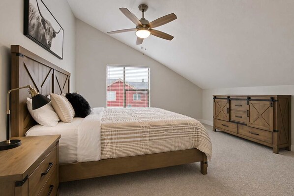 A bedroom with a bed, a nightstand, a desk, a wardrobe, a ceiling fan, and a window with a view of a red building. - Alluring Farmhouse Styled Town Home (McCall)