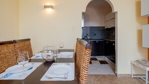 A dining area with a table set for two, a kitchen in the background, and a woven chair design. - Casa Hermes (Olbia)