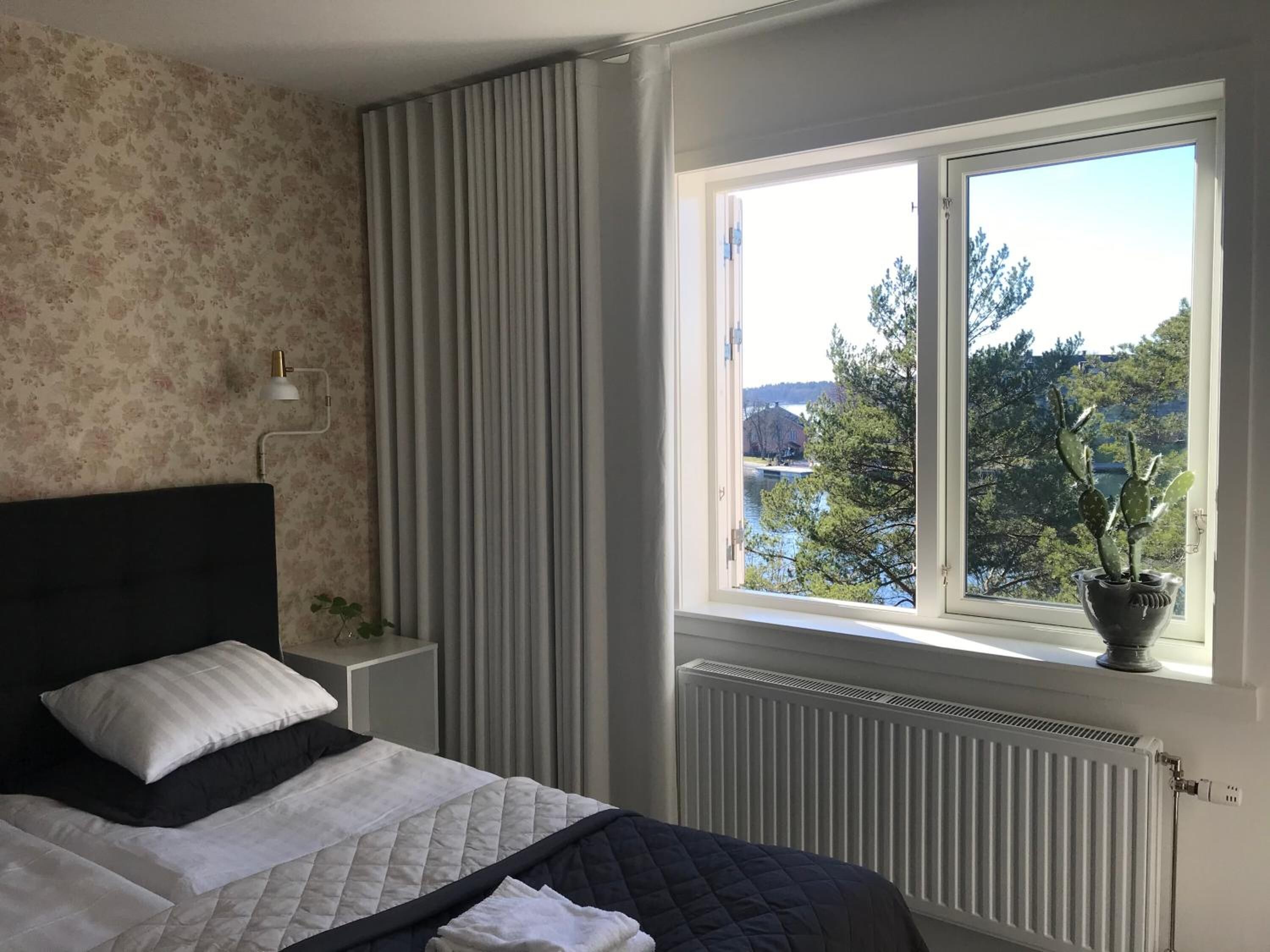 Een slaapkamer met een bed, nachtkastje, lamp, raam met uitzicht op een meer en bomen, en een radiator.