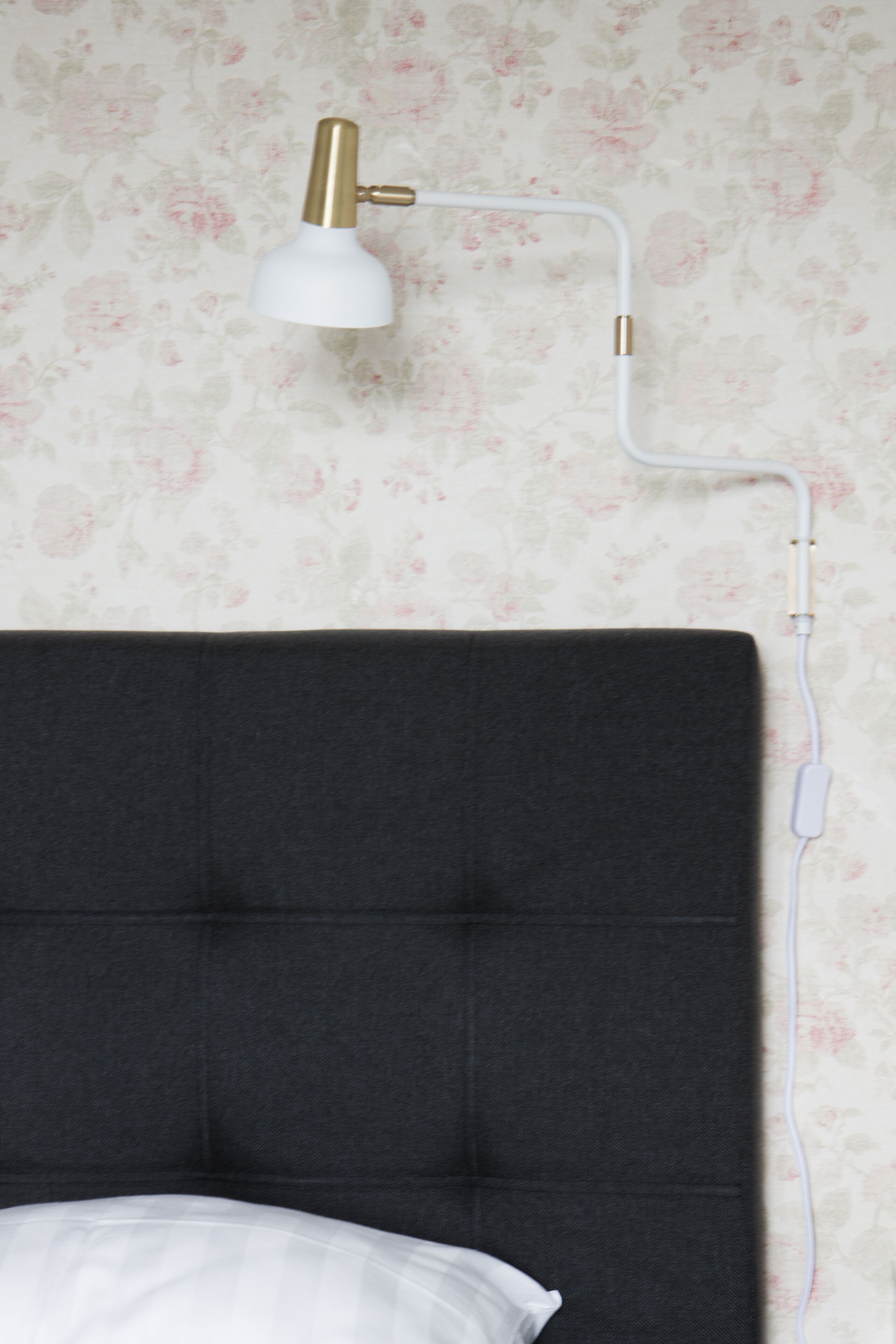 Een bed met een donker hoofdbord, een wit kussen en een muurlamp met een witte kap en een messing voet.