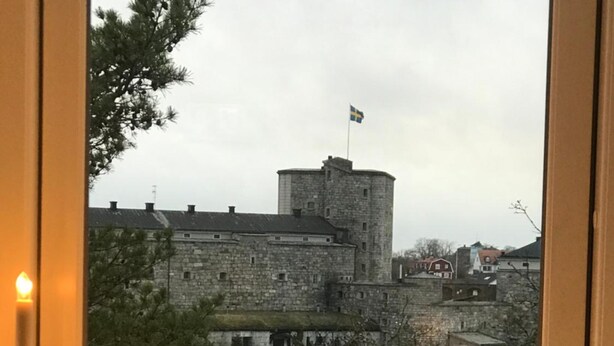 A view of a stone castle with a flag, a body of water, and bare trees.
